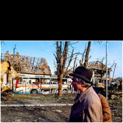 A civilian walks past the wreckage of a destroyed city bus amid heavily damaged buildings and shattered trees following intense shelling in Vukovar.
The scene reflects the scale of destruction in the city after the fighting, with public infrastructure reduced to ruins and everyday urban life abruptly brought to a halt.

The photograph documents the civilian experience of war in the immediate aftermath of the siege, emphasizing the contrast between ordinary human presence and the devastation of the urban environment.