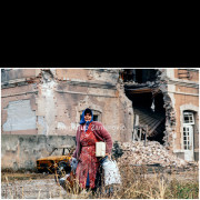 Vukovar (Croatia) – November 18, 1991

A civilian woman carries personal belongings while standing in front of a heavily damaged residential building in Vukovar following the end of major combat operations.
The destroyed facade, collapsed interior structures, scattered debris, and abandoned vehicle in the background illustrate the extent of devastation suffered by civilian neighborhoods during the siege of the city.

The photograph documents the immediate civilian experience in the aftermath of urban warfare, capturing displacement and survival amid widespread destruction.