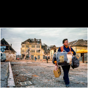 Vukovar (Croatia) – November 1991

A civilian woman walks through the devastated streets of Vukovar carrying personal belongings salvaged from her destroyed home.
Behind her, heavily damaged buildings with collapsed roofs and shattered façades testify to the scale of destruction following months of intense fighting.

The photograph documents the civilian experience of war and displacement during the final days of the siege of Vukovar.