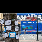 Passing through Pančevo on February 10, 2019, I encountered a temporary stage prepared for a political rally titled “Future of Serbia.” The visual contrast between the official stage design — dominated by a large blue LED screen displaying an idyllic tropical landscape — and the improvised obituary notices taped to a tree in the foreground creates a striking and unintended commentary on the political and social moment.

The image shown on the stage, photographed somewhere in the Pacific, suggests a distant promise of paradise, while the surrounding urban space tells a far more complex story. The obituary notices, placed there independently of the event, align almost symbolically with the rally’s title, transforming the scene into a layered visual metaphor of contemporary political reality.