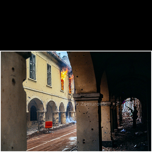 Vukovar (Croatia) – November 1991
A Serb fighter moves through the arcaded street of Vukovar as a building burns above the damaged colonnade.
The historic architecture is heavily scarred by shelling, with fire still visible inside upper-floor rooms, illustrating the intensity of urban combat during the final phase of the siege.
The photograph documents armed presence and destruction within the old town area.