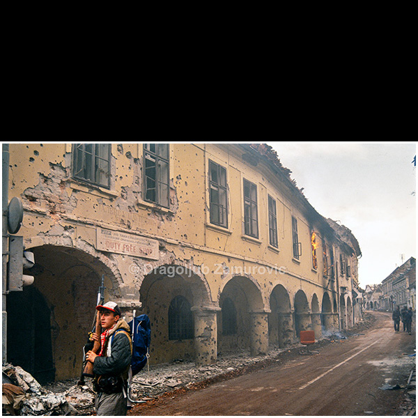 Vukovar (Croatia) – November 1991
A Serb fighter walks along a heavily damaged street in Vukovar, passing arcaded buildings scarred by shelling and small-arms fire.
The remains of urban infrastructure and ongoing fires inside the buildings reflect the intensity of combat during the final days of the siege.
The photograph documents armed movement through the devastated historic center of the town.