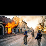 Vukovar (Croatia) – November 18, 1991

Serbian fighters move along a street in Vukovar as a residential building burns following heavy shelling during the final phase of the siege.
Flames, smoke, and damaged facades along the street illustrate the scale of destruction in the urban center after months of combat.

The photograph documents the immediate post-combat environment and the presence of armed participants amid widespread urban devastation.