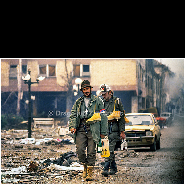 Vukovar (Croatia) – November 18, 1991
Serbian fighters walk through a heavily damaged urban area of Vukovar carrying supplies and personal items following the end of major combat operations.
Destroyed buildings, scattered debris, and abandoned vehicles in the background illustrate the extent of urban devastation after months of fighting.
The photograph documents the immediate post-combat environment and the everyday actions of armed participants amid the ruins of the city.