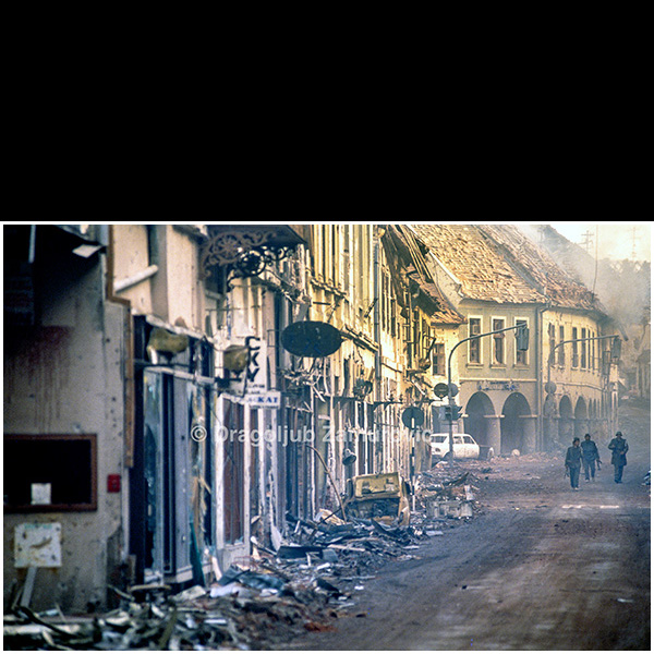 Vukovar (Croatia) – November 18, 1991
Serbian soldiers walk along a heavily damaged street in Vukovar following the end of major combat operations.
Shattered shopfronts, collapsed facades, debris-filled sidewalks, and damaged vehicles line the street, illustrating the extent of destruction inflicted on the historic city center during the siege.
The photograph documents the immediate aftermath of urban warfare, capturing the altered urban landscape and the presence of military personnel amid the ruins of civilian infrastructure.
