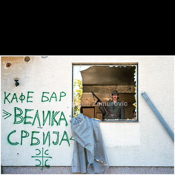 This was the first time in my life that I found myself in the midst of active warfare. I did not adapt easily, and relied heavily on advice from more experienced colleagues. Discipline among soldiers and volunteers was fragile, alcohol was consumed excessively, and behavior on the front line often bordered on chaos.
I had spent one year in the army in 1974, where I had already witnessed irresponsibility, negligence, and various forms of absurdity, but I could not have imagined such conduct in an active war zone.
Slobodan Milošević, a member of the Communist Party since the age of seventeen, sent the Yugoslav People’s Army (JNA) and paramilitary volunteers to the front line in Croatia. At the same time, Franjo Tuđman, the youngest general under Tito, was shaping an ethnically homogeneous state. Radical nationalist movements promoted the idea of a “Greater Serbia,” yet in many of the areas where they operated, almost no Serbs remained.
The photograph shows an armed soldier standing guard inside a destroyed café bar, framed by a shattered window. The graffiti on the wall — “Café Bar Velika Srbija” — stands in stark contrast to the emptiness and destruction surrounding him, reflecting the grim reality of nationalist slogans colliding with everyday life on the front line.