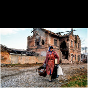 A civilian woman walks through the devastated streets of Vukovar, carrying personal belongings salvaged from her destroyed home. Behind her stands the heavily damaged Vukovar railway station, its collapsed roof and shattered façade bearing witness to months of intense shelling and street fighting.
The photograph documents the civilian experience of war and displacement during the final days of the siege of Vukovar, capturing a moment of quiet endurance amid widespread destruction.