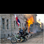 As Yugoslav People’s Army (JNA) forces and various paramilitary units enter Vukovar, armed men ride through devastated streets while buildings burn in the background. The photograph captures a moment of chaos and contradiction: military presence, destruction, and gestures of triumph amid a city still under fire. Taken during the final phase of the Battle of Vukovar, the image reflects the collapse of civilian life and the fragmentation of authority in the immediate aftermath of combat.