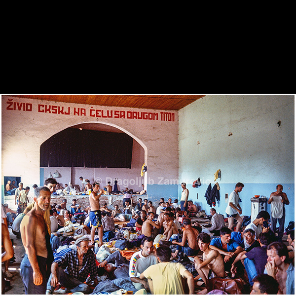 This photograph was taken in August 1992 during an organized visit by journalists to detention camps in Manjača, Omarska, and Trnopolje. The visit was approved by Sonja Karadžić, then Head of the Cabinet of the Ministry of Information of the Republika Srpska. Journalists were transported by bus from one camp to another, in an apparent attempt to demonstrate that detainees were not uniformly emaciated, following the international publication of photographs showing severely malnourished prisoners from Trnopolje.
The image shows a crowded interior space where detainees are gathered with their personal belongings, reflecting the conditions inside the camp at the time. While individual experiences varied, many of the men held in these camps endured severe physical and psychological suffering. This photograph documents the visible reality encountered during that visit, serving as a historical record of the environment and circumstances within the Trnopolje camp.