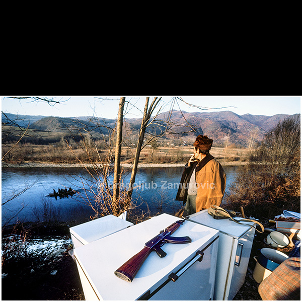 In January 1993, during the war in Bosnia and Herzegovina, large numbers of refugees crossed the Drina River from Bosnia into Serbia. Reports of this mass movement brought me to the riverbank near the village of Crvice, where I encountered scenes that exceeded any prior expectations.
People were leaving their homes carrying only basic belongings, crossing the river in improvised boats, often in harsh winter conditions. Among the refugees were men, women, and elderly people. One striking detail was the presence of firearms — not hunting rifles, but military weapons — carried by some of those fleeing.
This photograph captures a moment on the riverbank: improvised luggage, a rifle placed on household appliances, and a man looking toward the river as a boat with refugees reaches the opposite shore. The image stands as a documentary record of one of many, rarely documented episodes of wartime displacement during the winter of 1993.